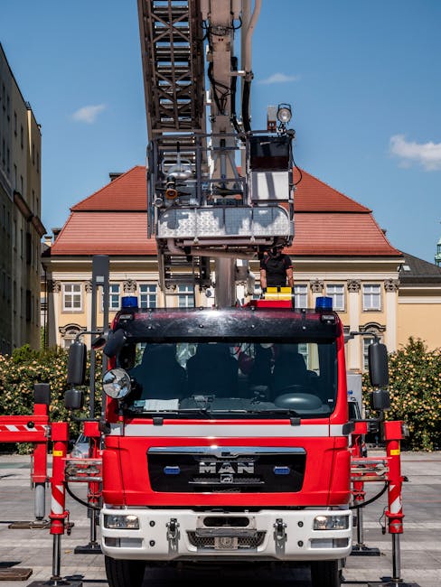 The image shows a red maintenance truck equipped with a tall extendable ladder or cherry picker, positioned outdoors on a paved area. The ladder extends vertically towards a building with classic arch